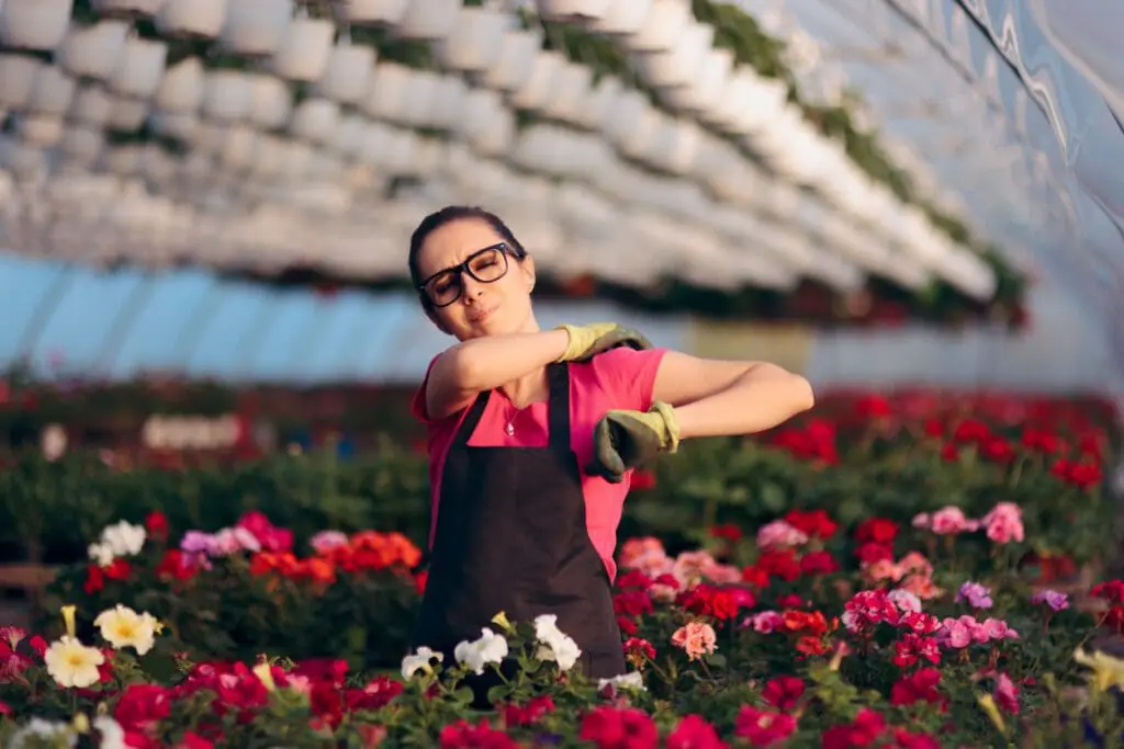 A woman stretching her shoulder in a greenhouse. First aid for gardeners.